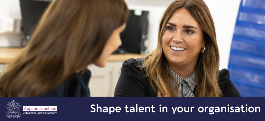 A female senior teacher smiles at a teaching apprentice as she sits with her at a desk to host a meeting. She is smiling at the apprentice who is talking to her and has her back to the camera.